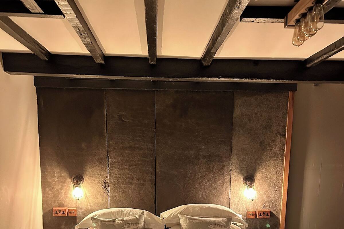 A neatly made bed with white linens and multiple pillows, flanked by two bedside tables with lamps, in a room with exposed wooden beams and a textured wall.