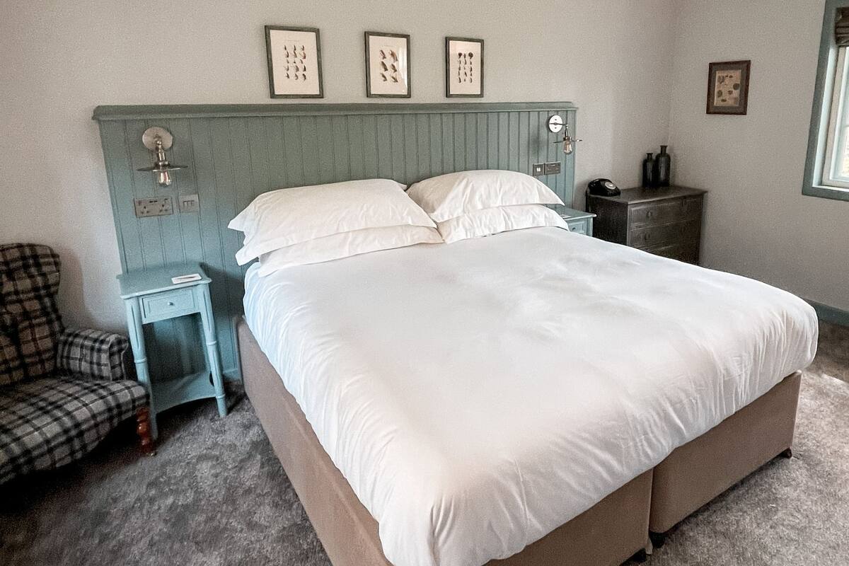 A bedroom with a large bed, a chair, a nightstand, and a dresser. There are framed pictures on the wall and a window with a view of trees.