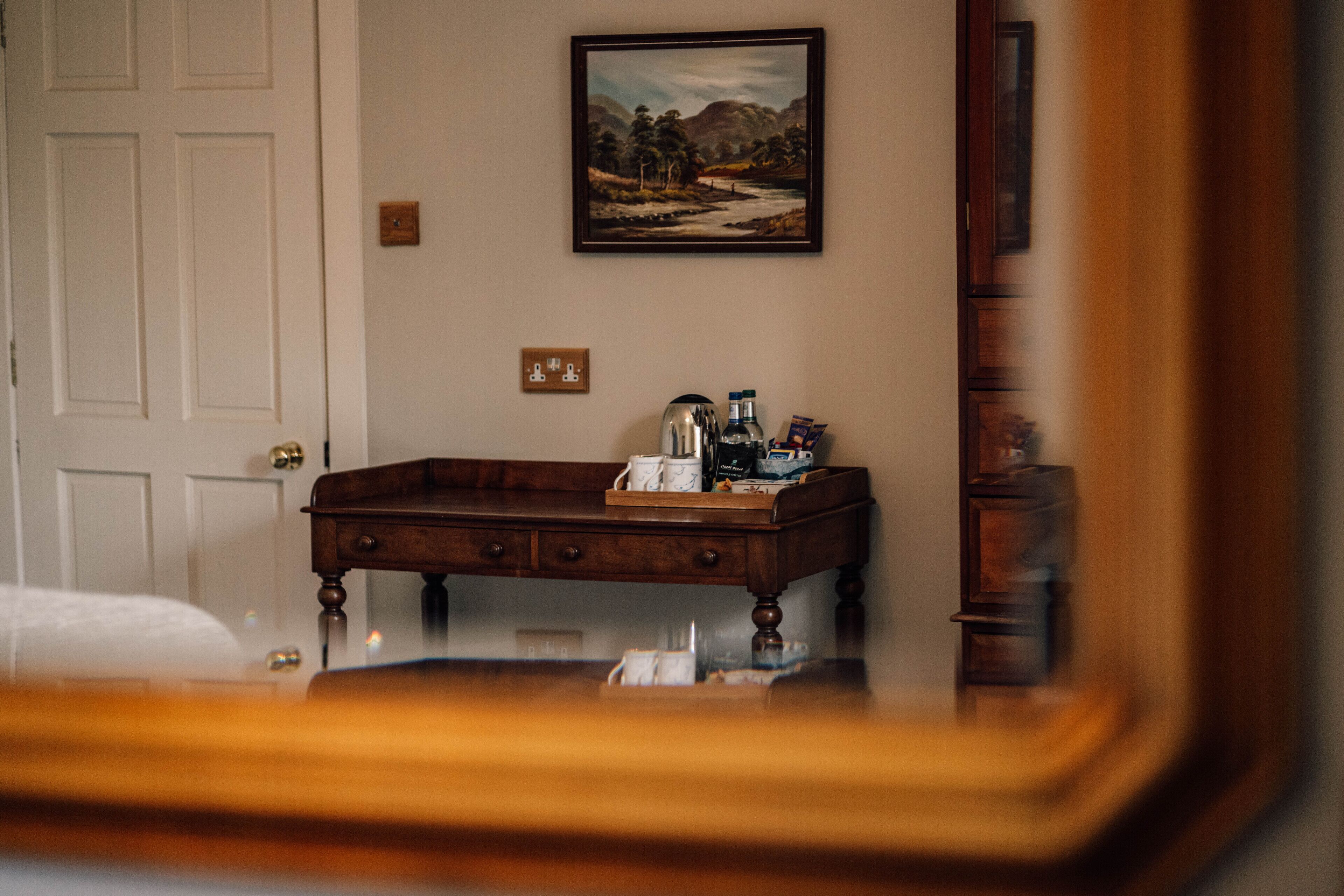Une chambre d’hôtel avec un bureau en bois, un tableau accroché au mur et un miroir qui reflète une partie de la pièce.