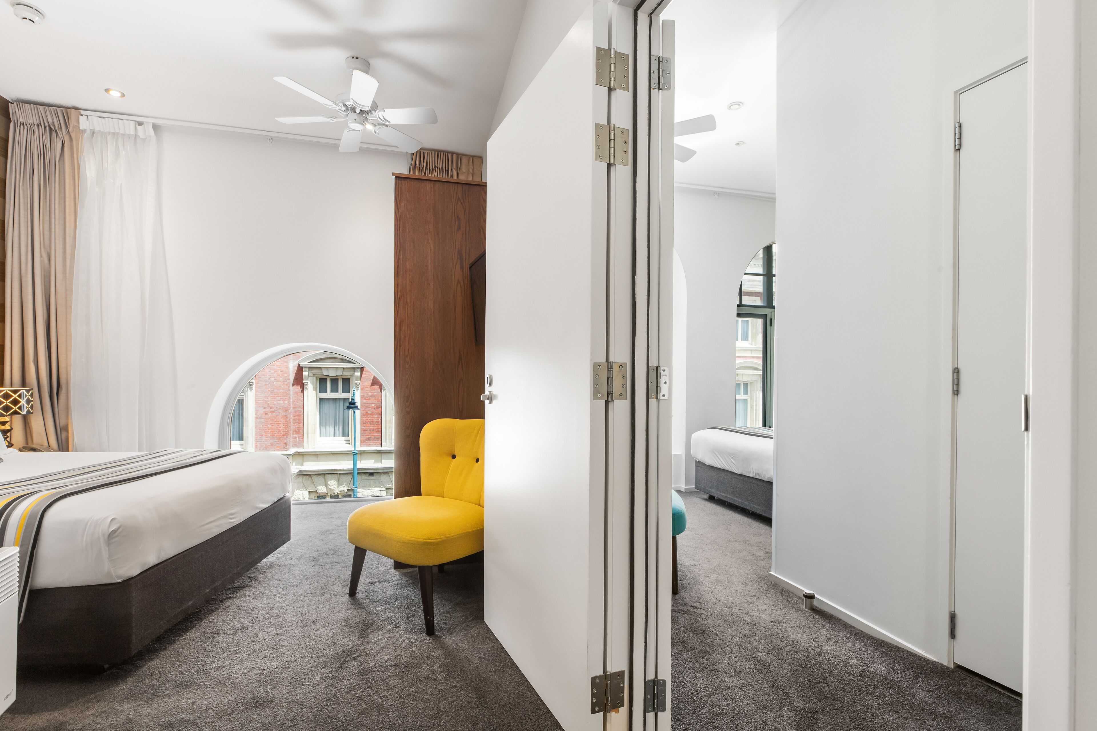 Una habitación de hotel con cama, una silla amarilla, un armario y vista a un edificio a través de una ventana.