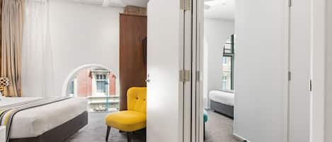 Una habitación de hotel con cama, una silla amarilla, un armario y vista a un edificio a través de una ventana.