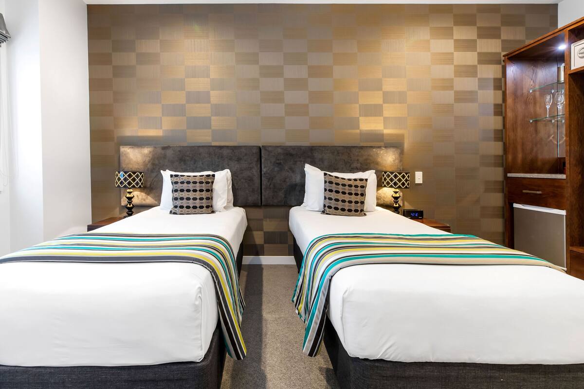 Une chambre d’hôtel avec deux lits, un motif à carreaux sur le mur, des tables de chevet avec des lampes et une armoire en bois.