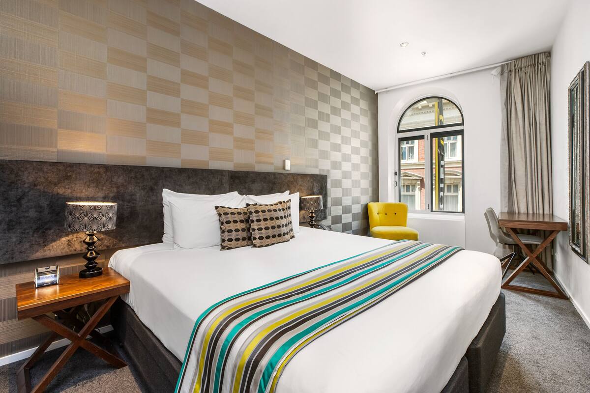 Une chambre d’hôtel avec un grand lit, une table de chevet avec une lampe, une chaise, un bureau et une fenêtre avec des rideaux.