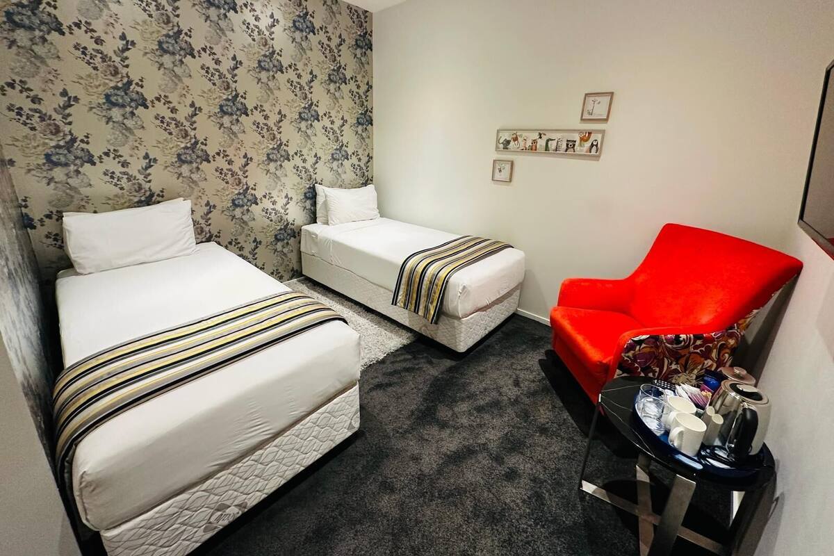 Une chambre d’hôtel avec deux lits, un fauteuil rouge, une petite table sur laquelle se trouve un plateau avec divers objets, et un papier peint à motifs floraux.