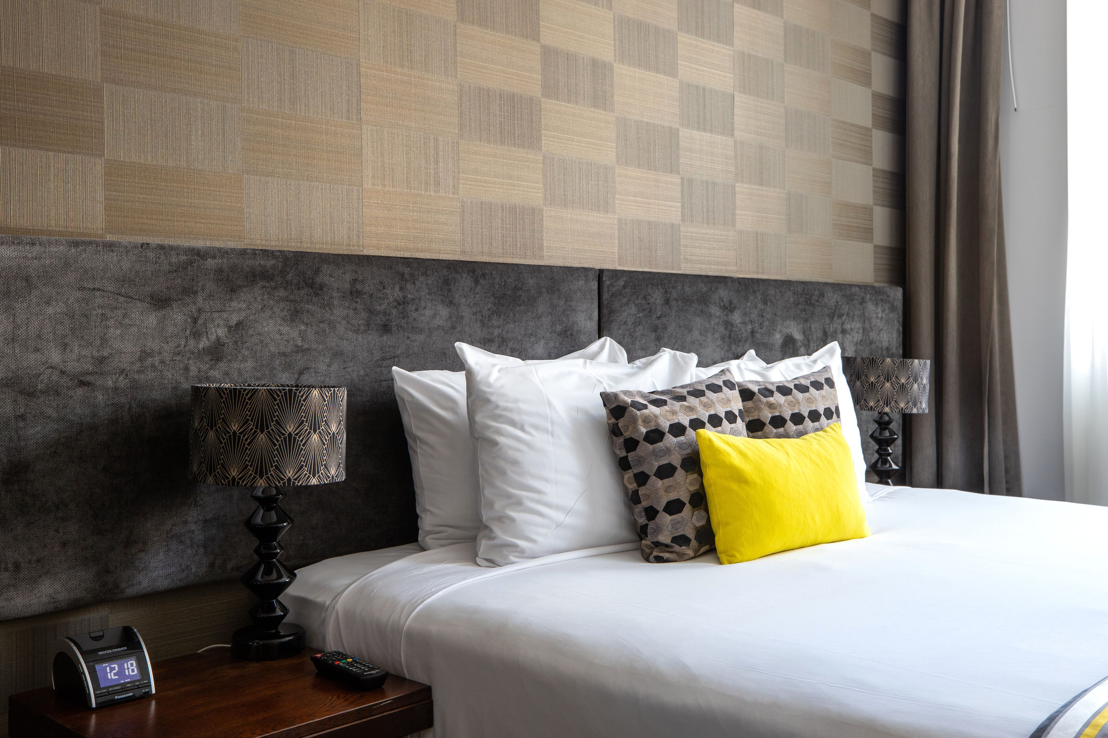 Una cama bien hecha con almohadas blancas y estampadas, un reloj digital y una lámpara en una mesita de noche de madera.
