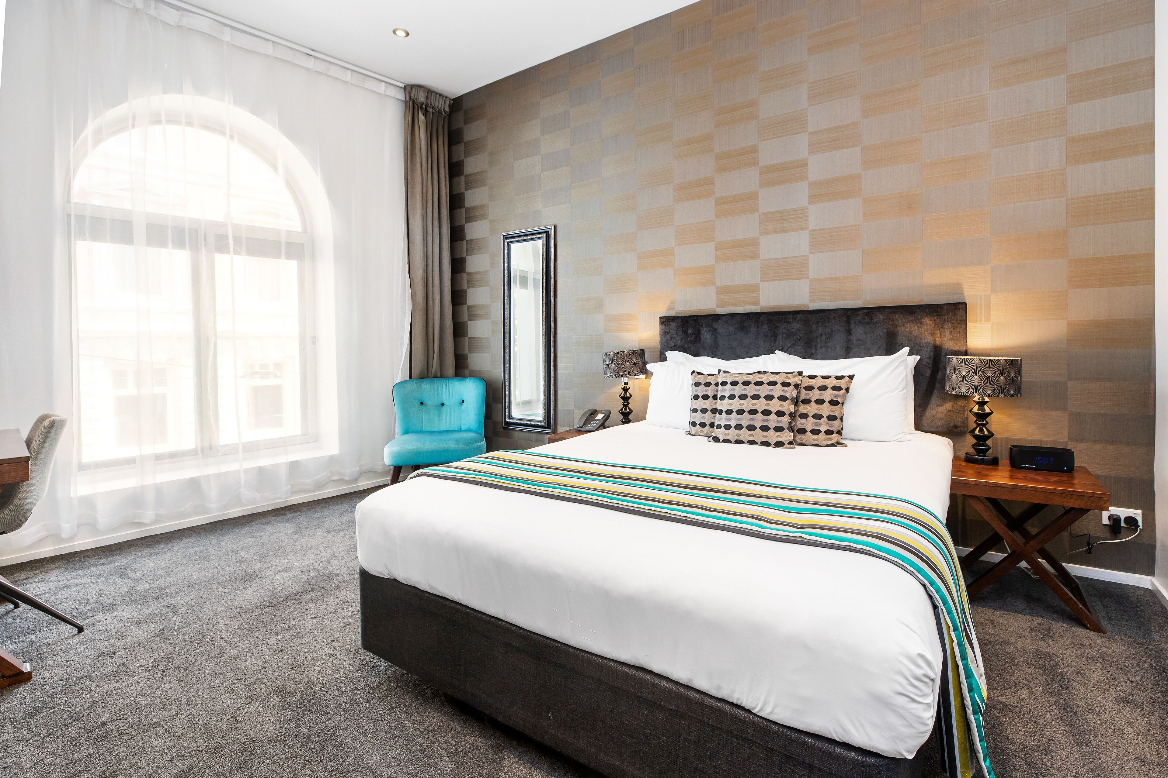 A hotel room with a large bed, a nightstand with a lamp, a chair, and a large window with curtains.