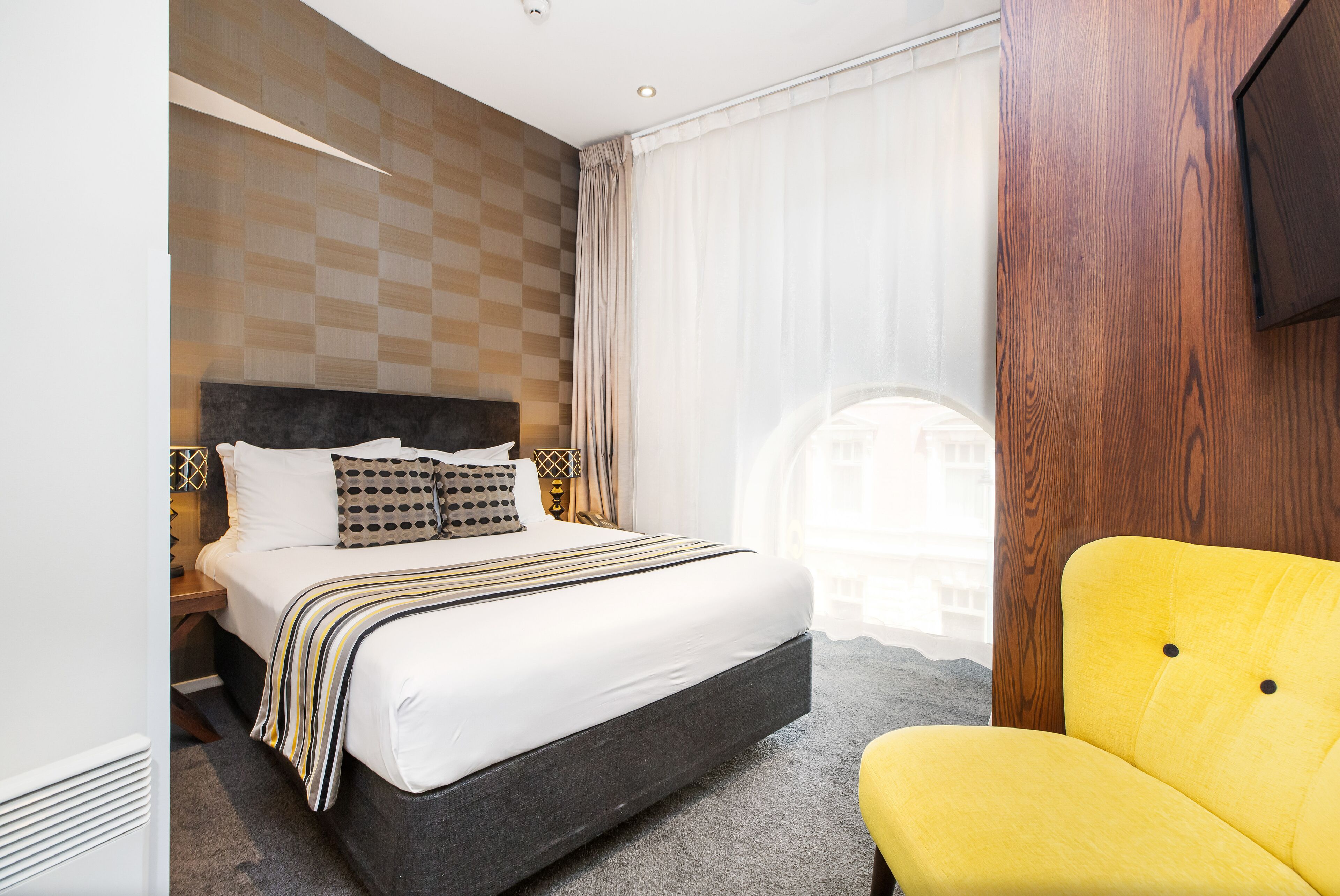 A hotel room with a large bed, a wooden headboard, a yellow armchair, and a view of a building through sheer curtains.