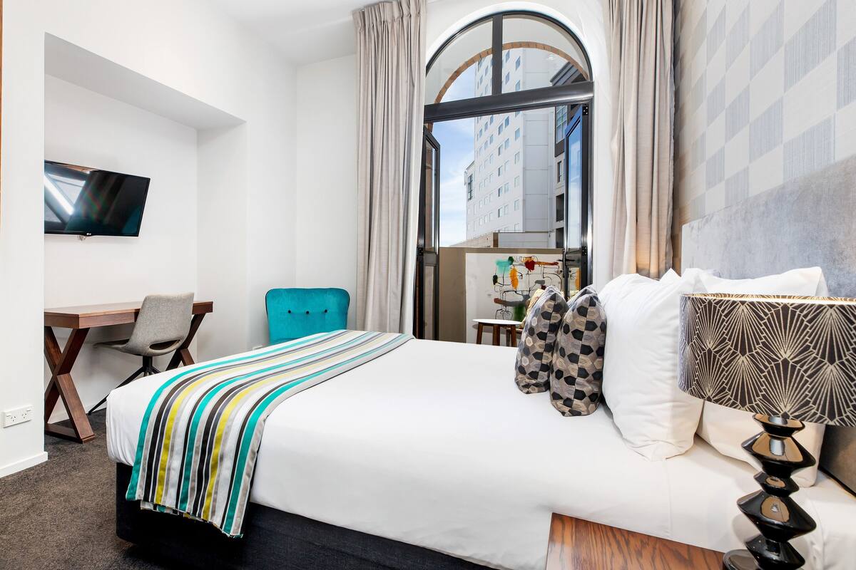 Une chambre d’hôtel équipée d’un lit, d’un bureau, d’une chaise, d’une télévision et offrant une vue sur les bâtiments par le biais d’une grande fenêtre.