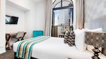 A hotel room with a bed, a desk, a chair, a TV, and a view of buildings through a large window.