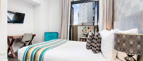 Habitación de hotel con cama, escritorio, silla, televisión y vistas a edificios a través de un ventanal grande.