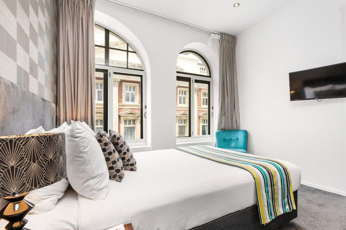 A hotel room with a bed, a TV, and large windows.