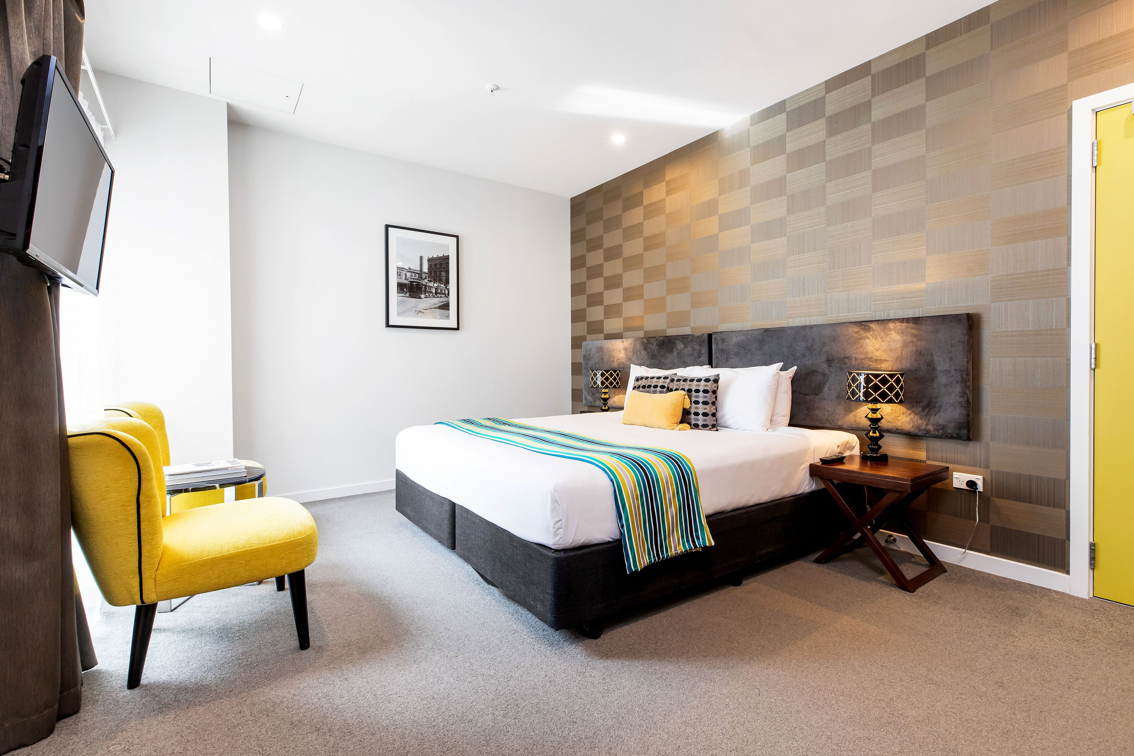 Una habitación de hotel moderna con una cama grande, una mesita de noche con lámpara, una silla y un televisor montado en la pared.
