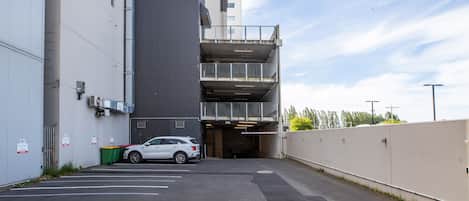 Entrada de un estacionamiento con un coche blanco dentro, un edificio de varios pisos con balcón, y cielo despejado.