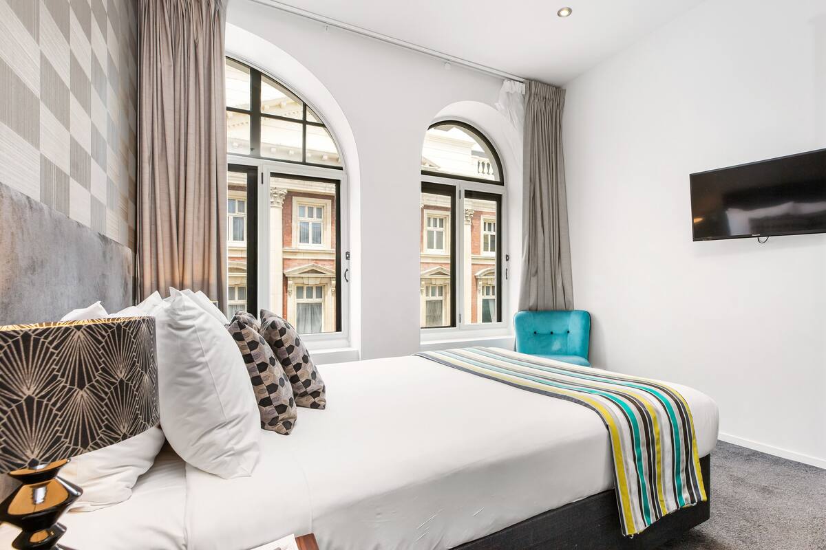 Une chambre d’hôtel moderne dotée d’un grand lit, d’un téléviseur fixé au mur et offrant une vue sur la ville grâce à de grandes fenêtres.