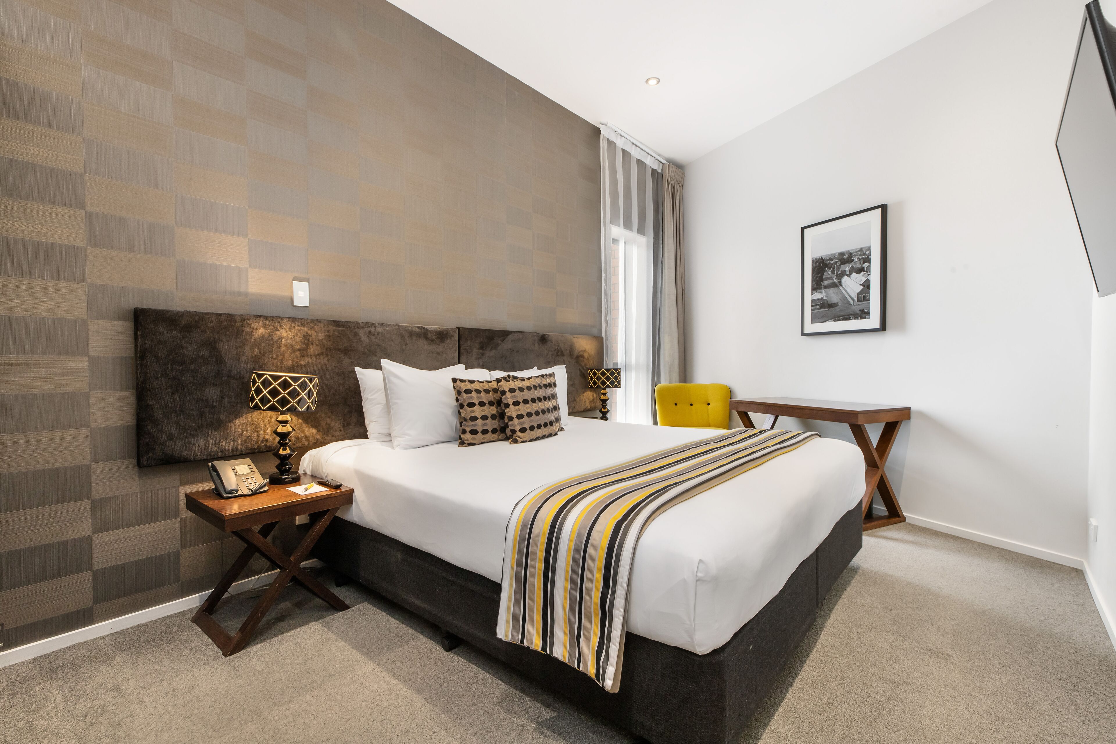 Habitación de hotel moderna con una cama grande, una mesita de noche con lámpara, una silla y un cuadro enmarcado en la pared.