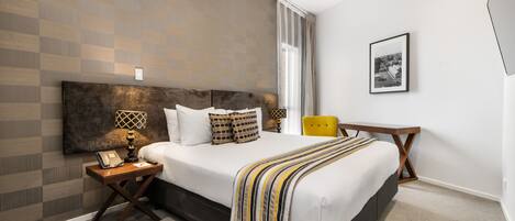 Habitación de hotel moderna con una cama grande, una mesita de noche con lámpara, una silla y un cuadro enmarcado en la pared.
