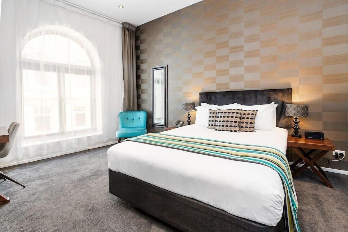 Une chambre d’hôtel avec un grand lit, un bureau, une chaise, une fenêtre avec des rideaux et un mur décoré d’un motif.
