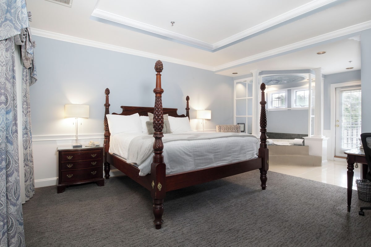 A four-poster bed with a wooden frame, a nightstand with drawers, a desk with a chair, and a large window with blinds.