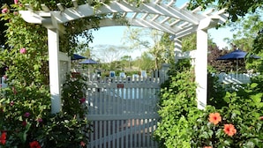 A gated entrance with a white picket fence, a pergola with climbing plants, and a brick pathway leading to a pool area.