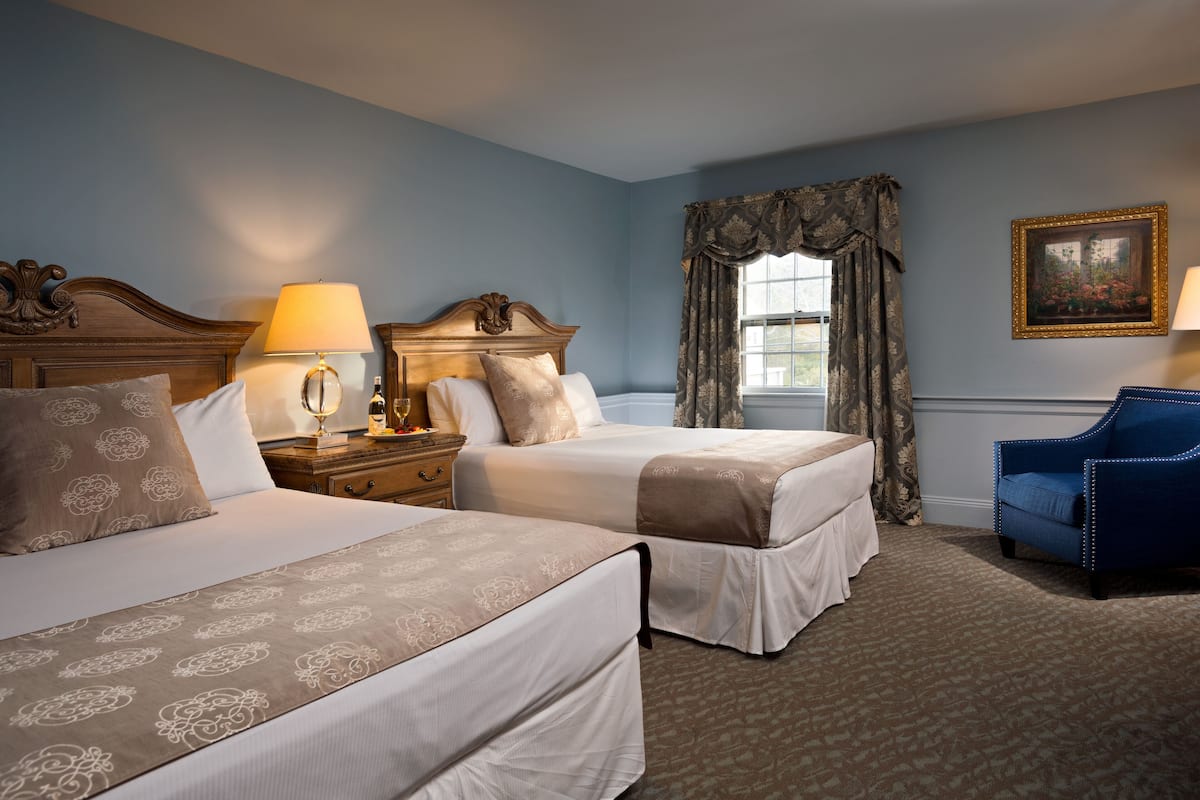 A hotel room with two beds, a nightstand with a lamp, a chair, and a window with curtains.