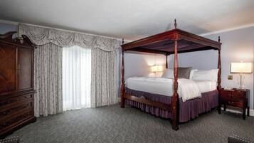 Una cama con dosel de madera y falda morada, una mesita de noche de madera con una lámpara, y un ventanal con cortinas de gasa.