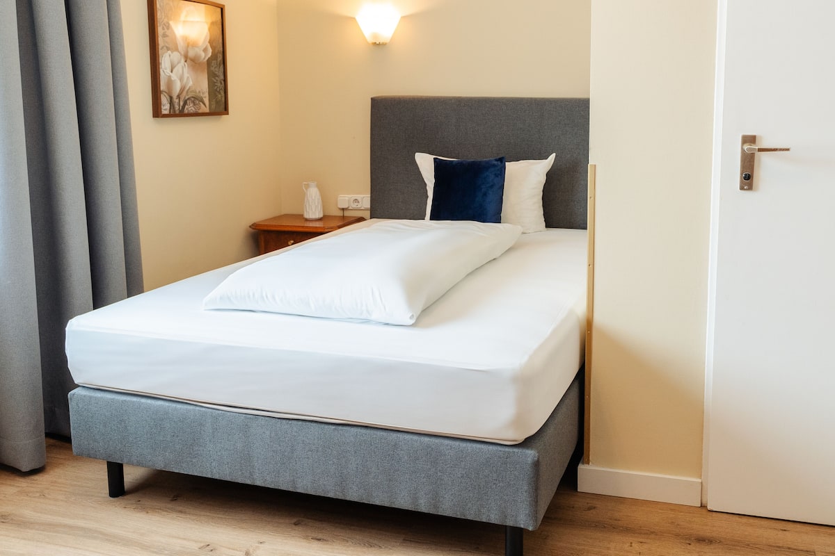 A neatly made bed with white linens, a grey headboard, and a bedside table with a lamp.