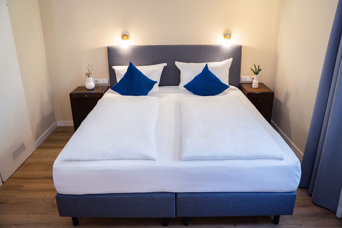 A neatly made bed with white bedding and blue pillows, flanked by two bedside tables with lamps and small plants.