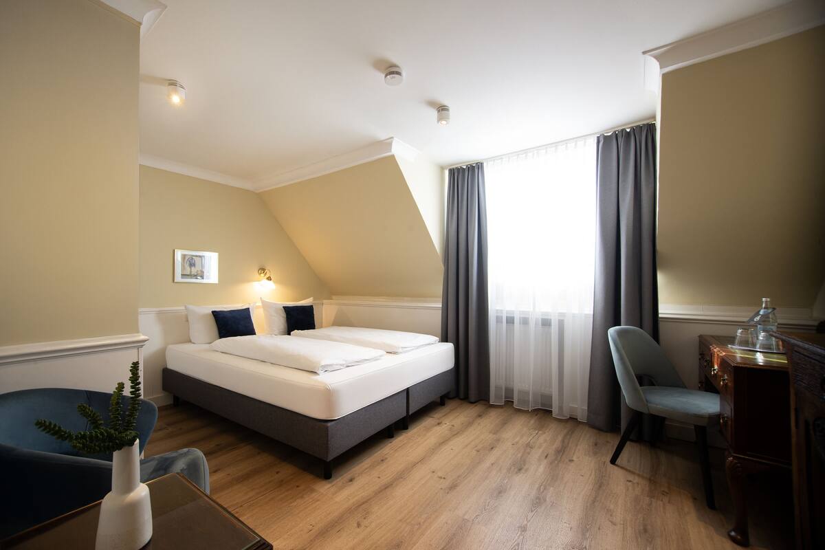 A hotel room with a bed, a desk, a chair, a plant, and a window with curtains.