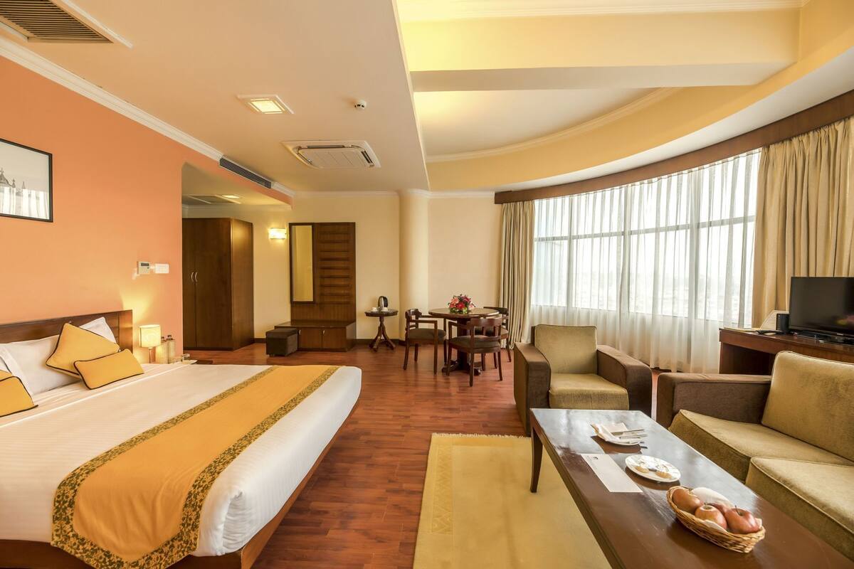 A hotel room with a large bed, a sofa, a dining table with chairs, a television, and a window with curtains.