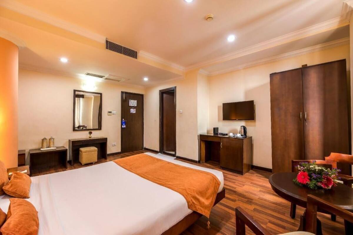 A hotel room with a bed, a desk, a chair, a table with flowers, a wardrobe, and a mirror.