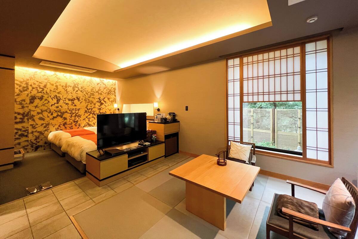 Ein modernes Hotelzimmer mit Flachbildfernseher, einem Holztisch, einer Couch und einem traditionellen japanischen Shoji-Fenster.