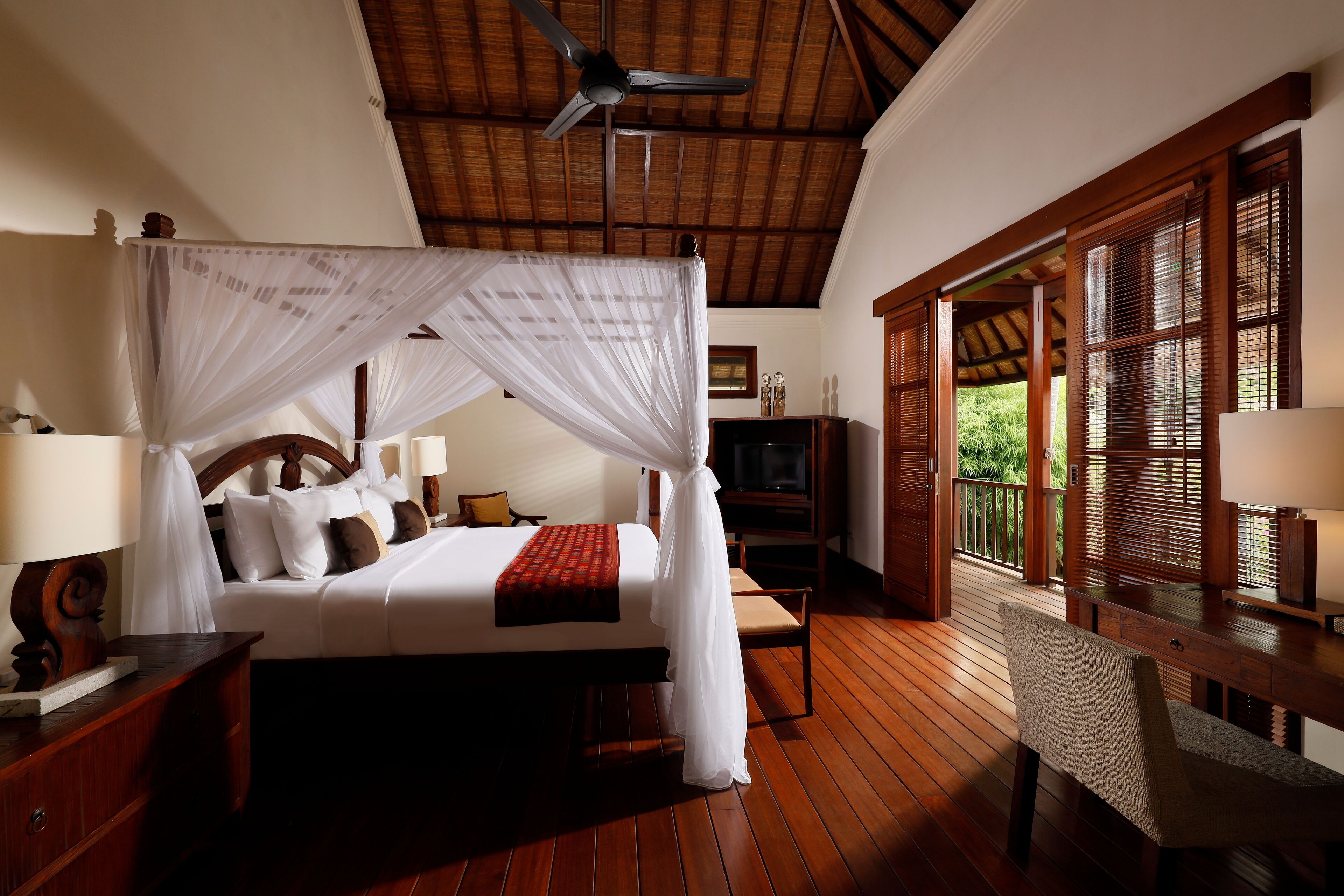 A spacious bedroom with a four-poster bed, wooden flooring, a ceiling fan, and a view of the outdoors through large windows.