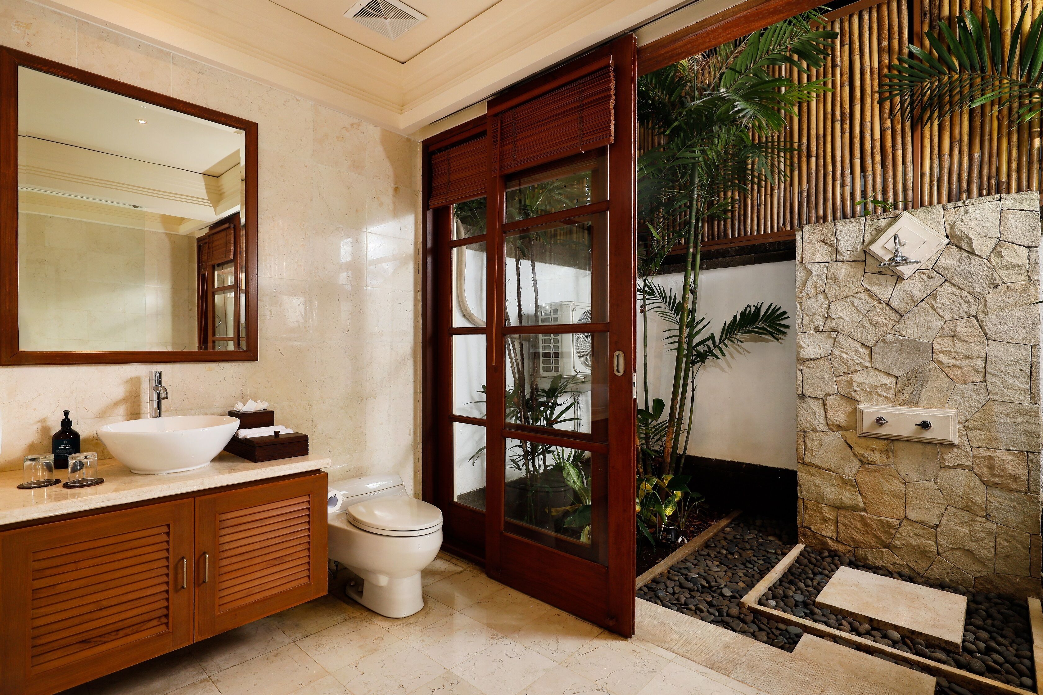 A bathroom with a large mirror, a white sink, wooden cabinets, a toilet, and a stone wall with a water feature.