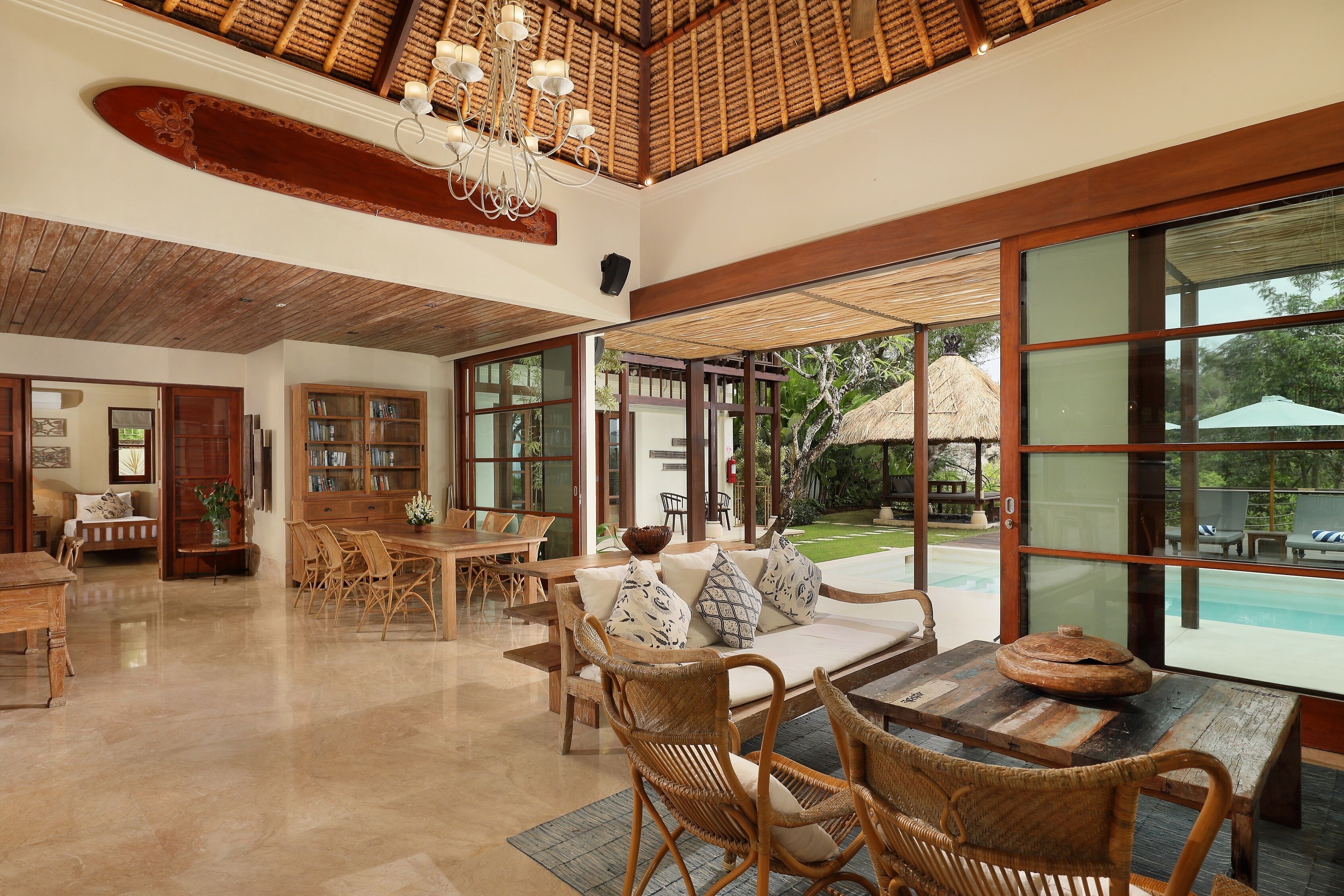 A spacious indoor area with a dining table, wicker chairs, a sofa with cushions, and a large window offering a view of an outdoor pool and garden.