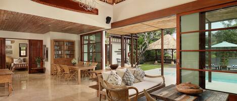 Un espacio interior amplio con una mesa de comedor, sillas de mimbre, un sofá con cojines y un ventanal con vistas a una piscina exterior y jardín.
