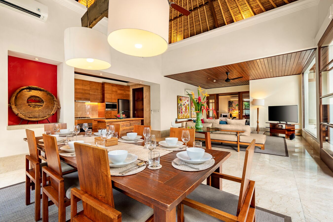 Uma sala de jantar espaçosa com uma mesa de madeira posta para seis pessoas, uma cozinha ao fundo, e uma sala de estar com televisão e assentos.