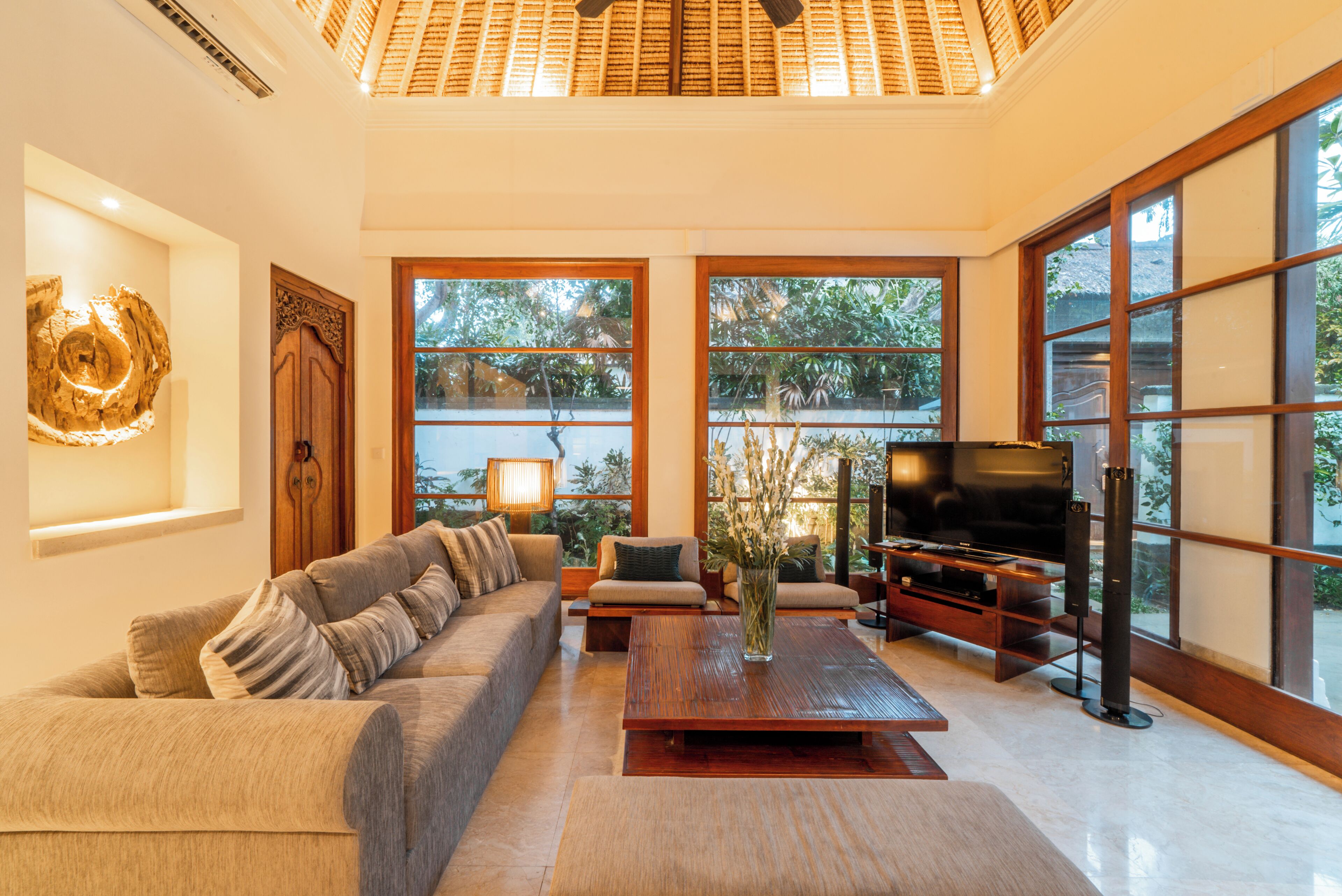 A spacious living room with a sofa, coffee table, and TV. Large windows provide natural light and views of greenery.