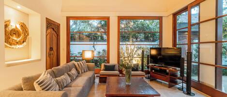 A spacious living room with a sofa, coffee table, and TV. Large windows provide natural light and views of greenery.