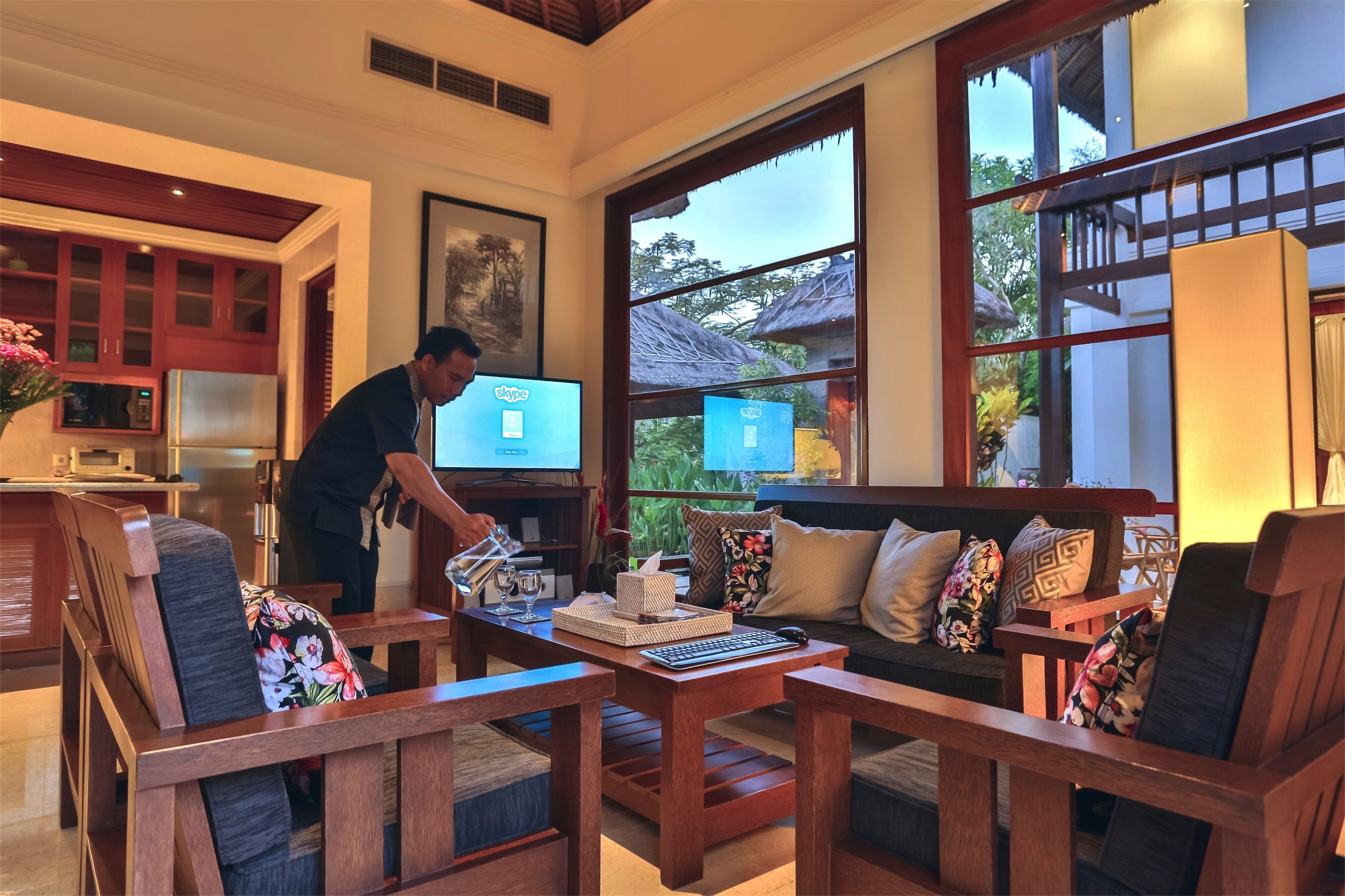 Un salón amplio con muebles de madera, un ventanal con vistas al exterior y una persona trabajando en un ordenador portátil.