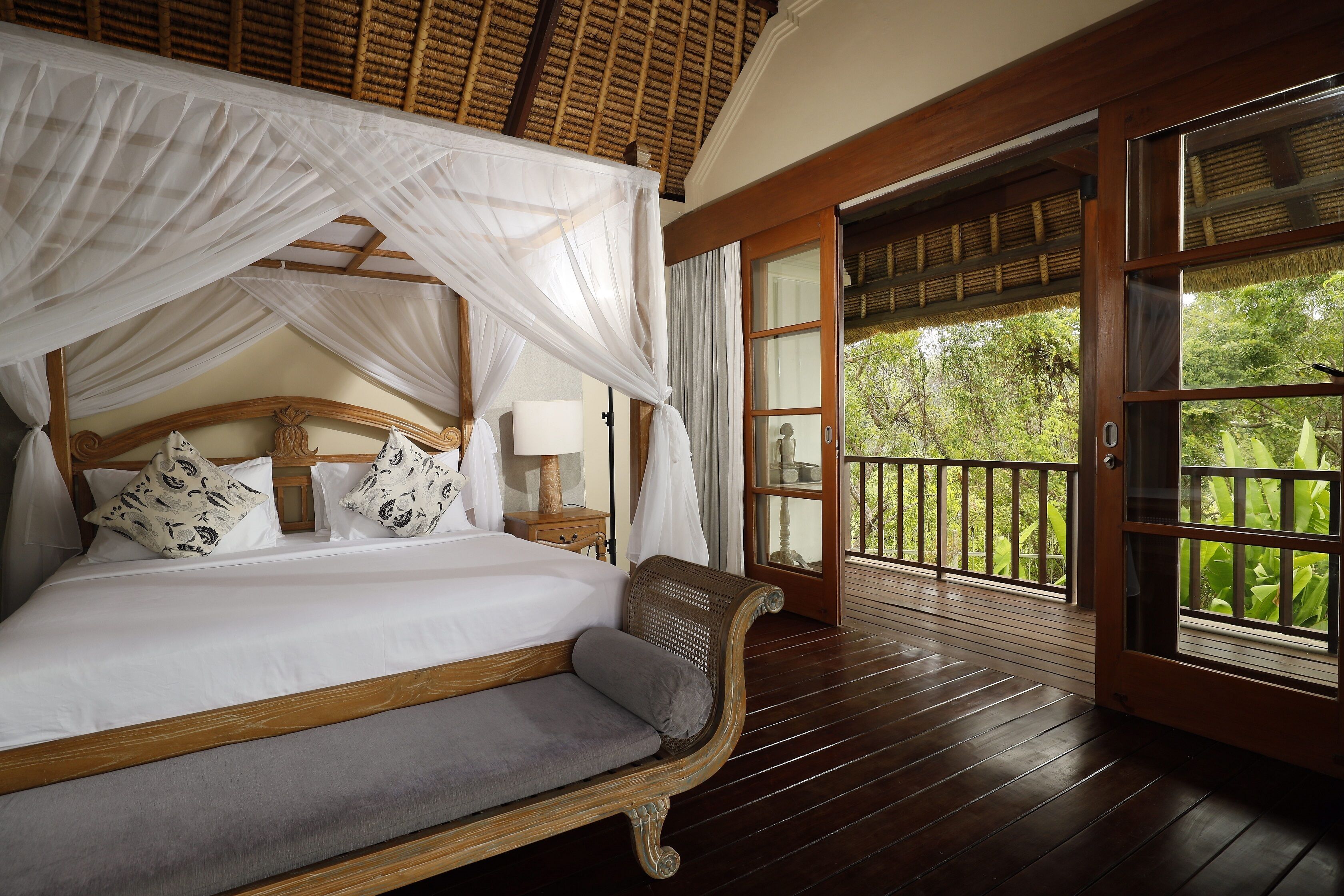 A bed with a canopy, a bench, and a view of greenery through a glass door.