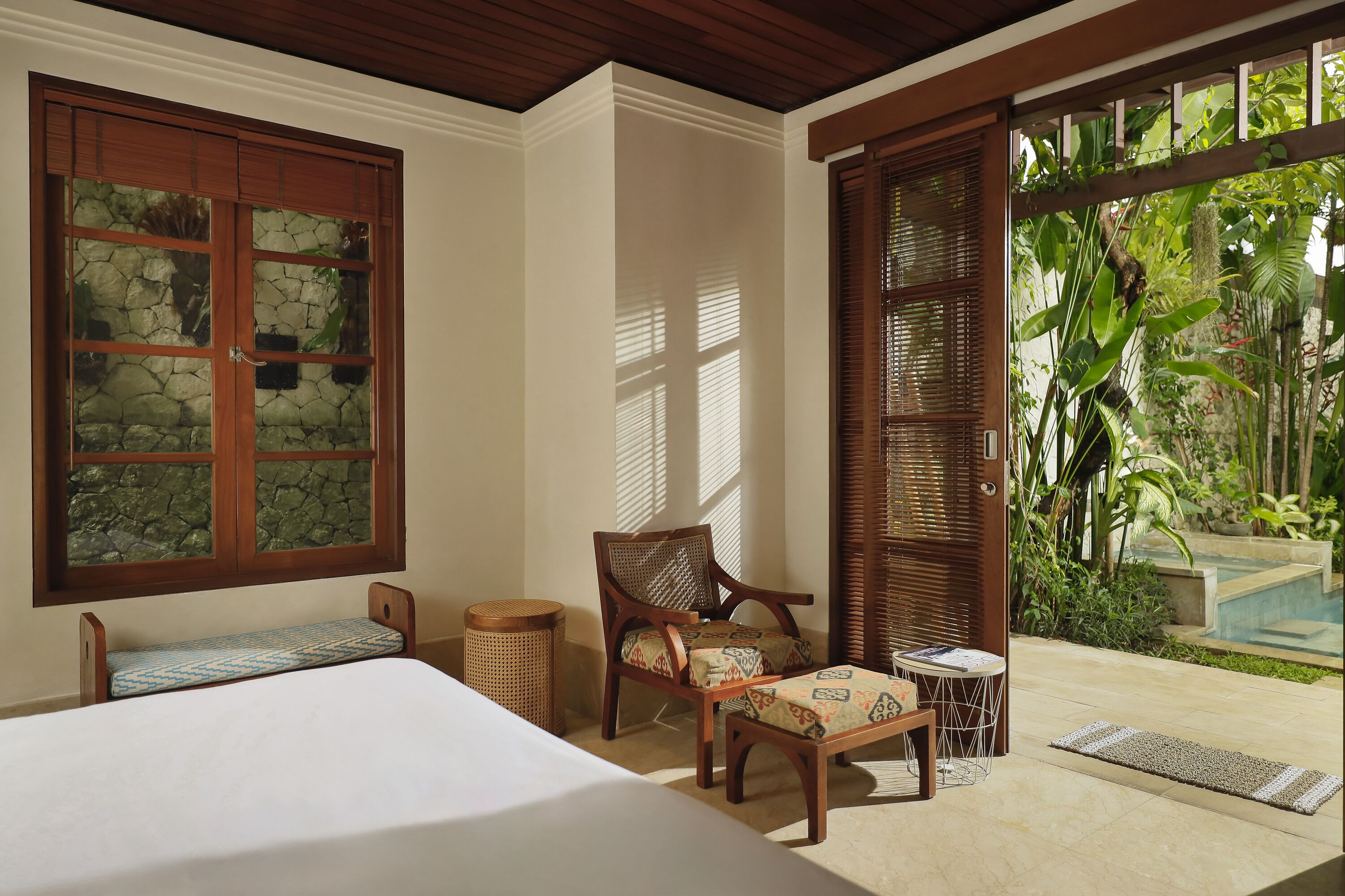 A room with a bed, a wooden chair, a small table, and a window with a stone wall view.