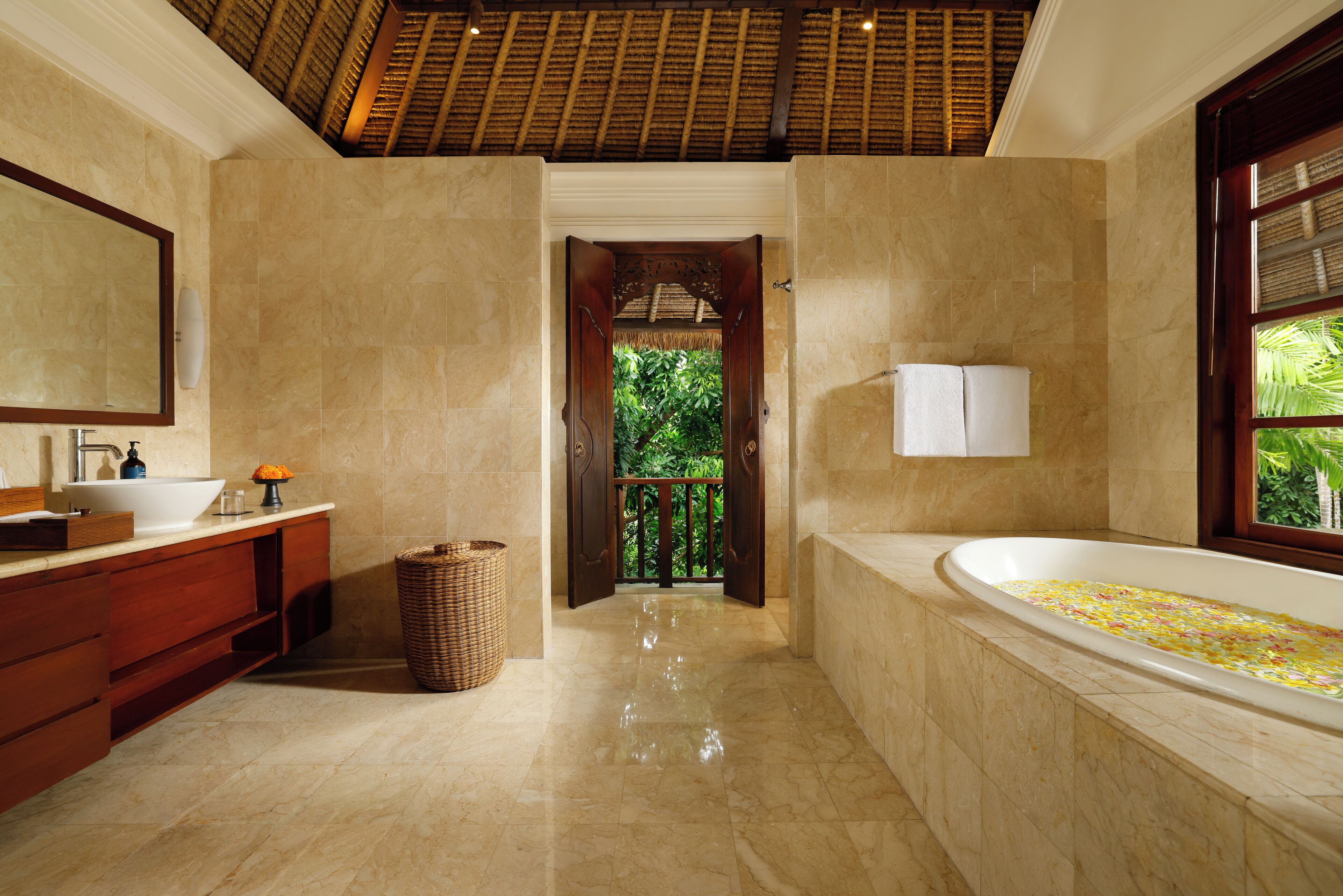 A bathroom with a large bathtub, a sink, and a mirror. There is a wooden door leading to a balcony with greenery outside.