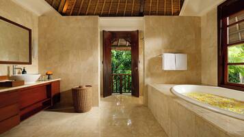 A bathroom with a large bathtub, a sink, and a mirror. There is a wooden door leading to a balcony with greenery outside.