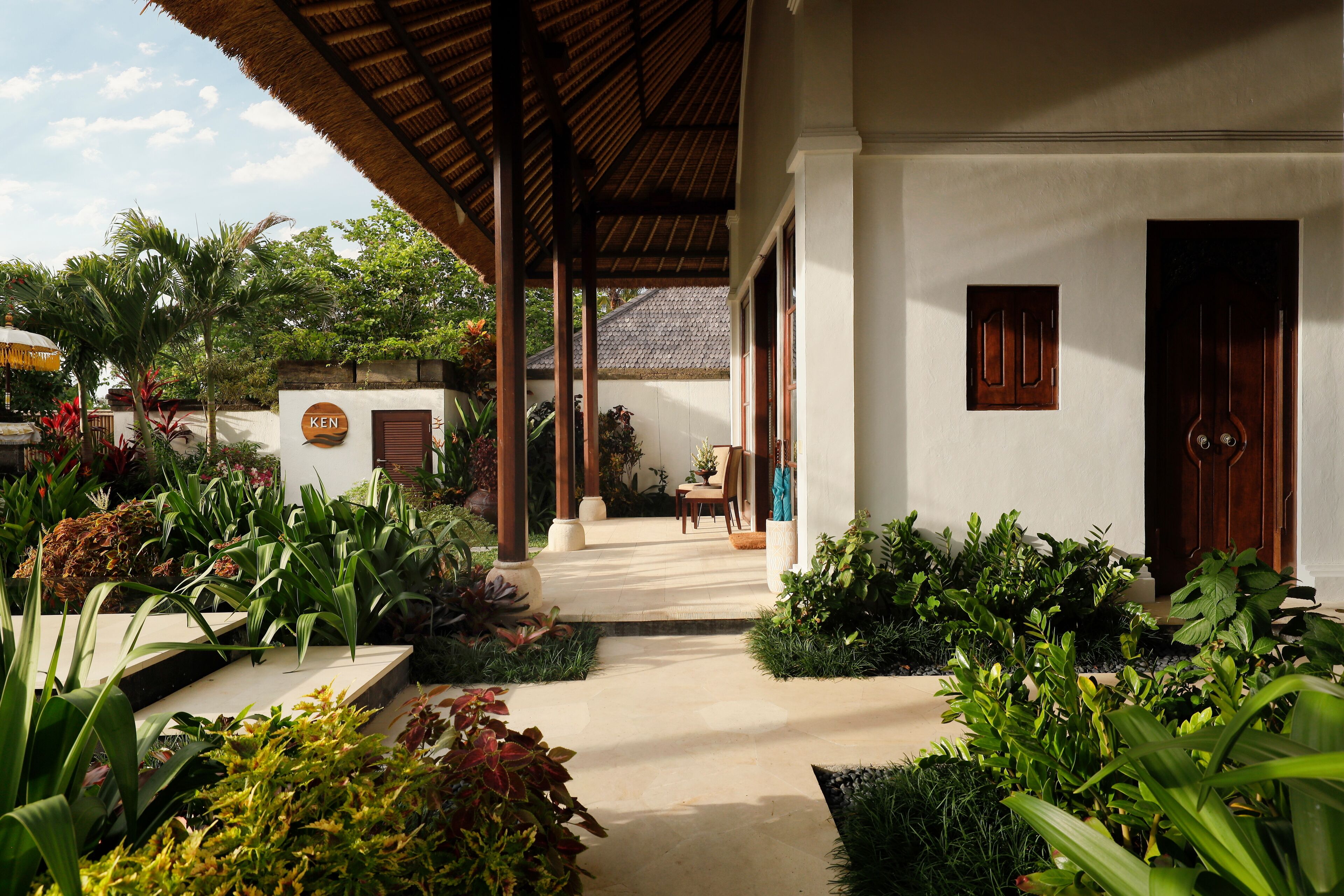 A covered outdoor area with a pathway leading to a building entrance, surrounded by lush greenery and a thatched roof structure.