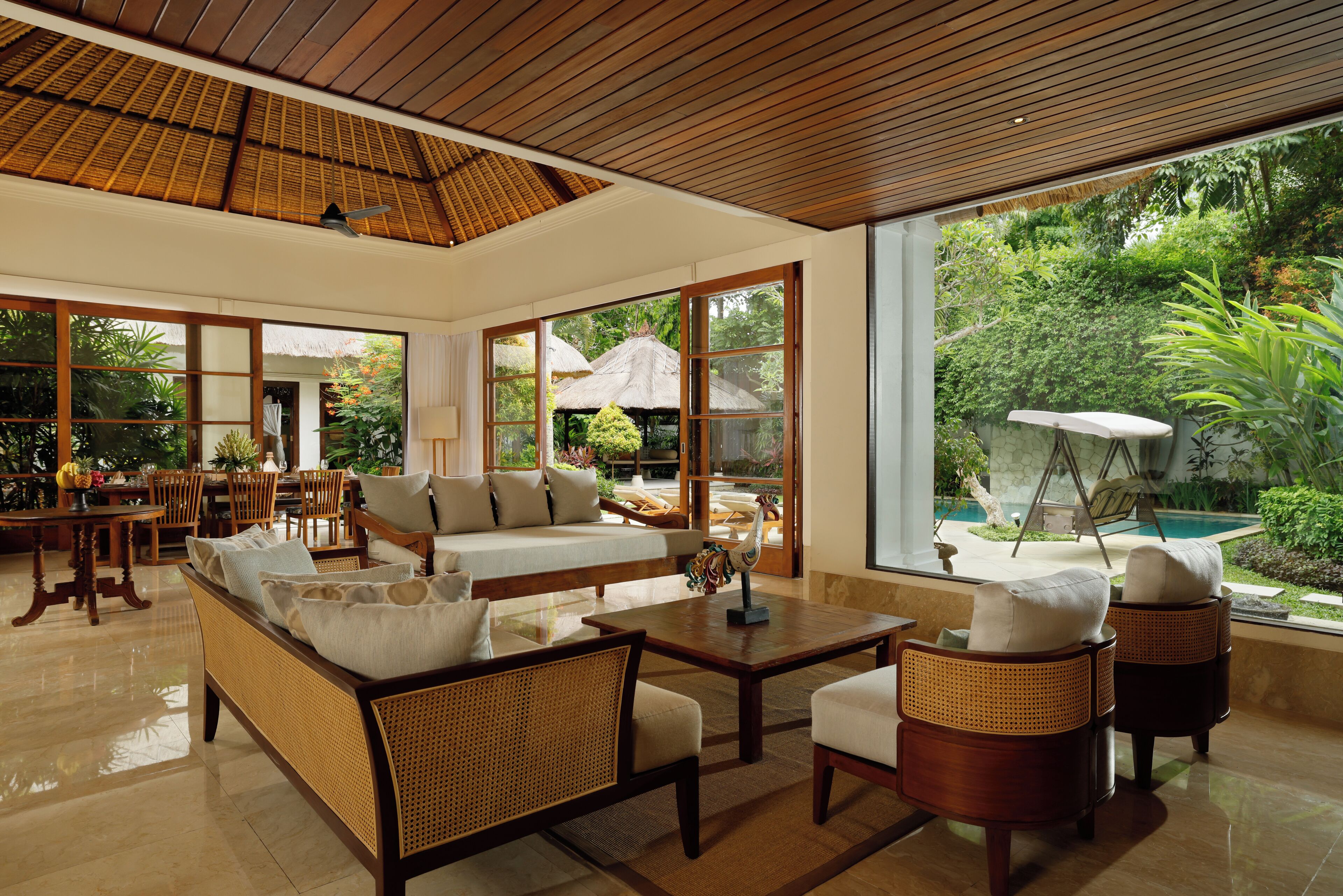 Amplia sala de estar con un sofá grande, dos sillones y una mesa de centro. Hay un comedor con una mesa y sillas, y se ve una piscina y abundante vegetación al exterior.