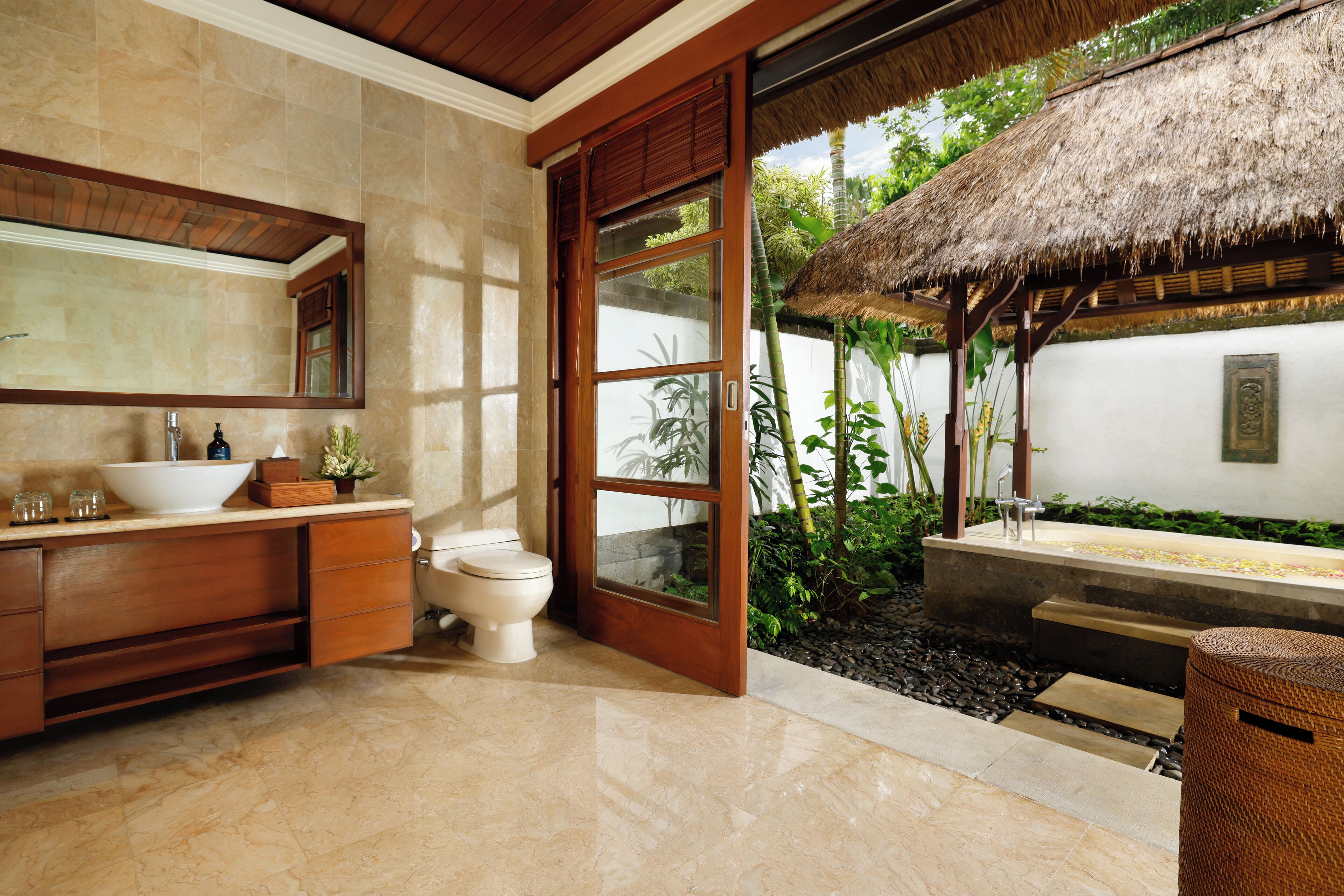 A bathroom with a large mirror, a white sink, and a toilet. There is a wooden door leading to a small outdoor area with a thatched roof and plants.