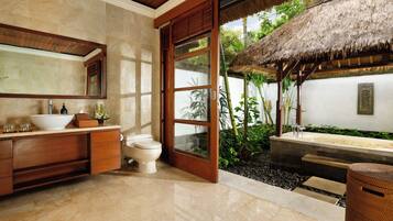 A bathroom with a large mirror, a white sink, and a toilet. There is a wooden door leading to a small outdoor area with a thatched roof and plants.