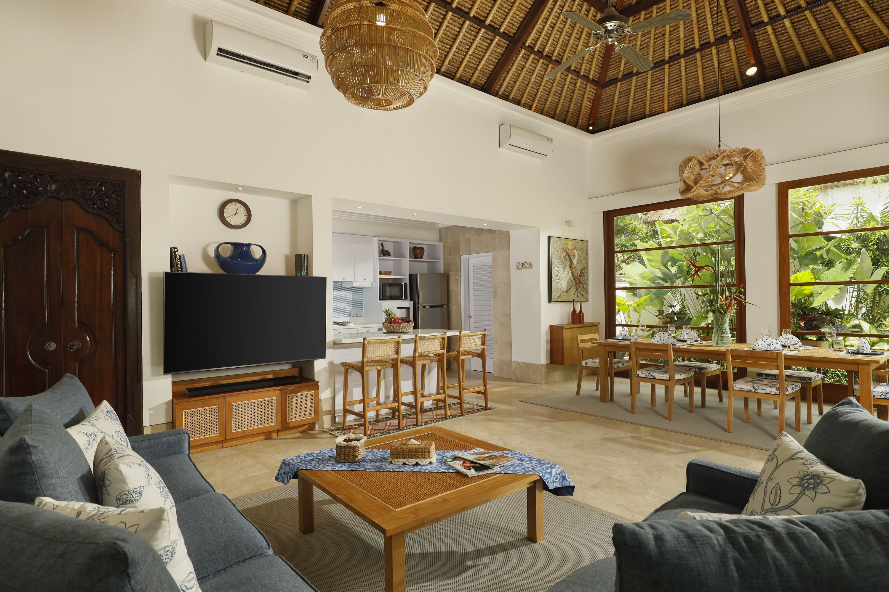 A spacious living room with a large flat-screen TV, a dining area with wooden chairs, and a kitchen in the background.