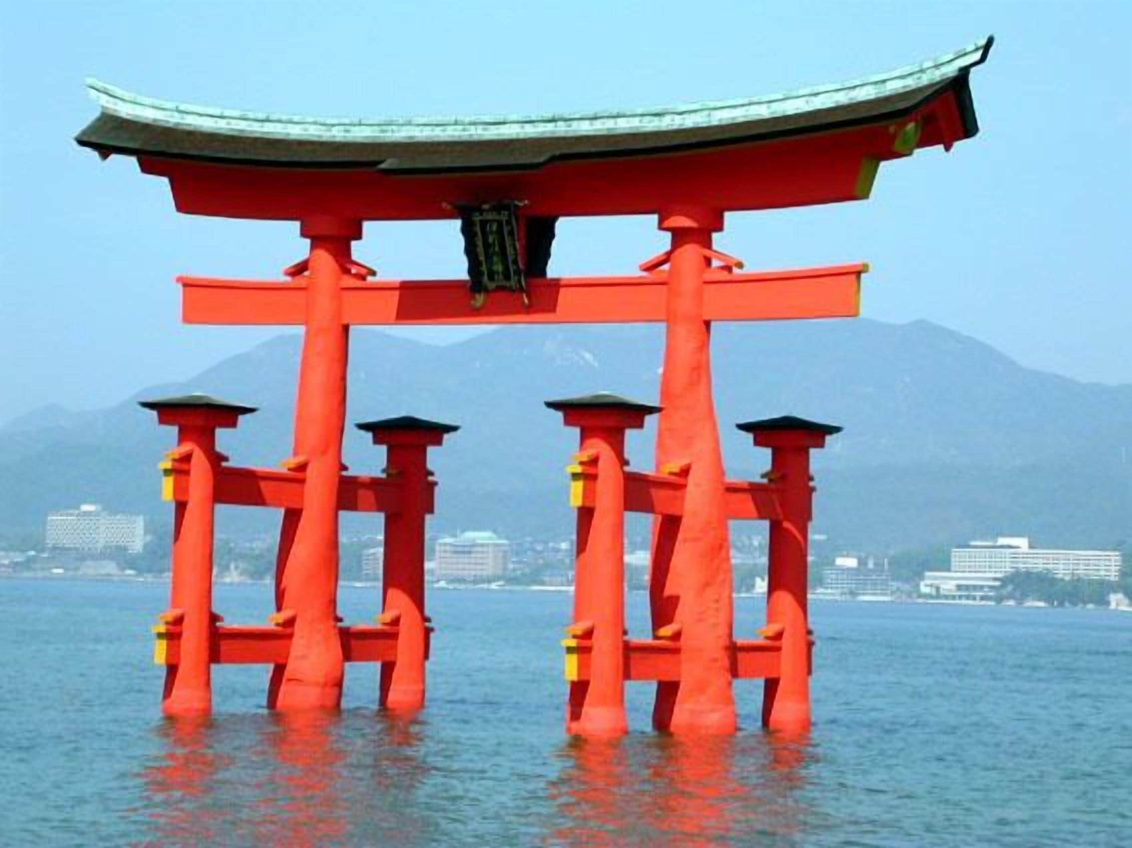 Un torii japonés tradicional de estructura roja y techo verde, situado en una masa de agua con montañas al fondo.