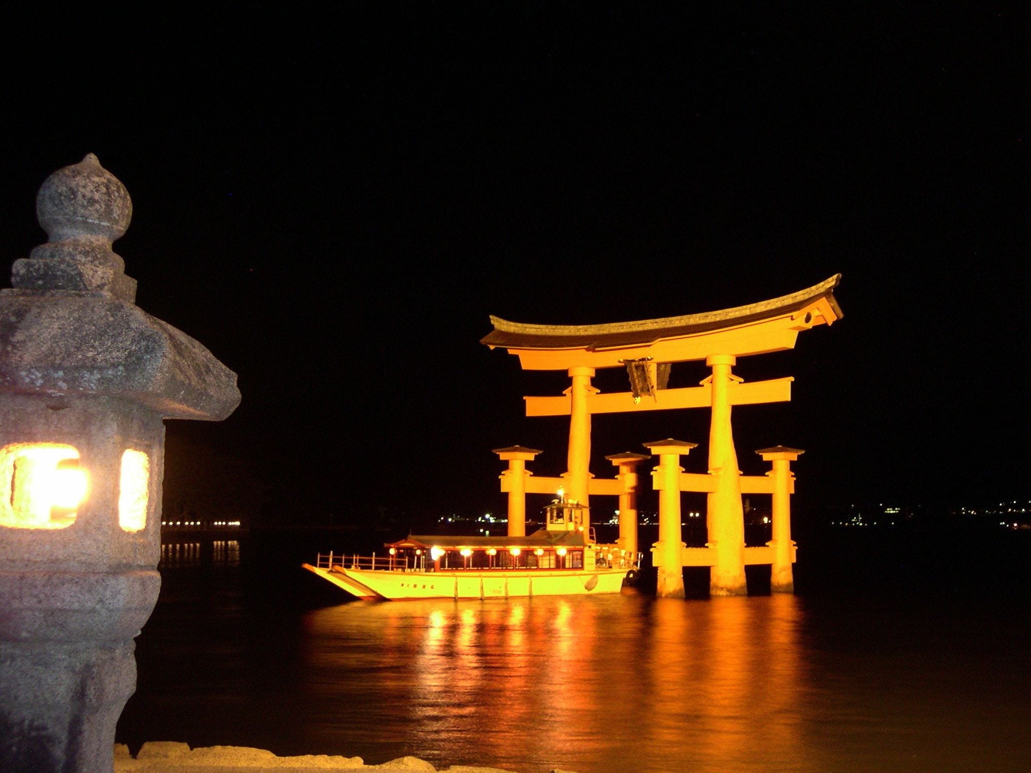 A traditional Japanese torii gate illuminated at night, with a stone lantern in the foreground and a boat docked nearby.